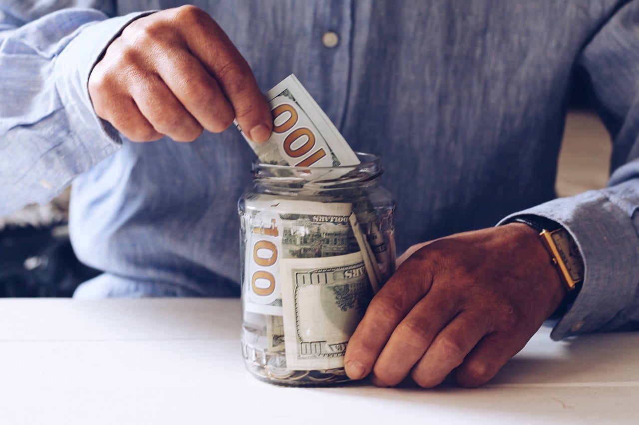 A person is shown putting a hundred dollar bill into a mason jar with other hundred dollar bills inside.