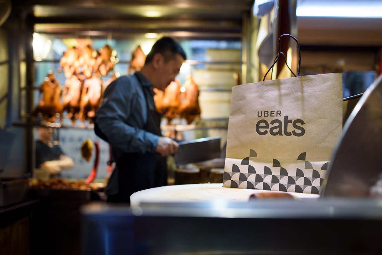 Medium shot of a paper food bag on a table while a chef cutting meat with a cleaver in background. (Photo courtesy Uber)