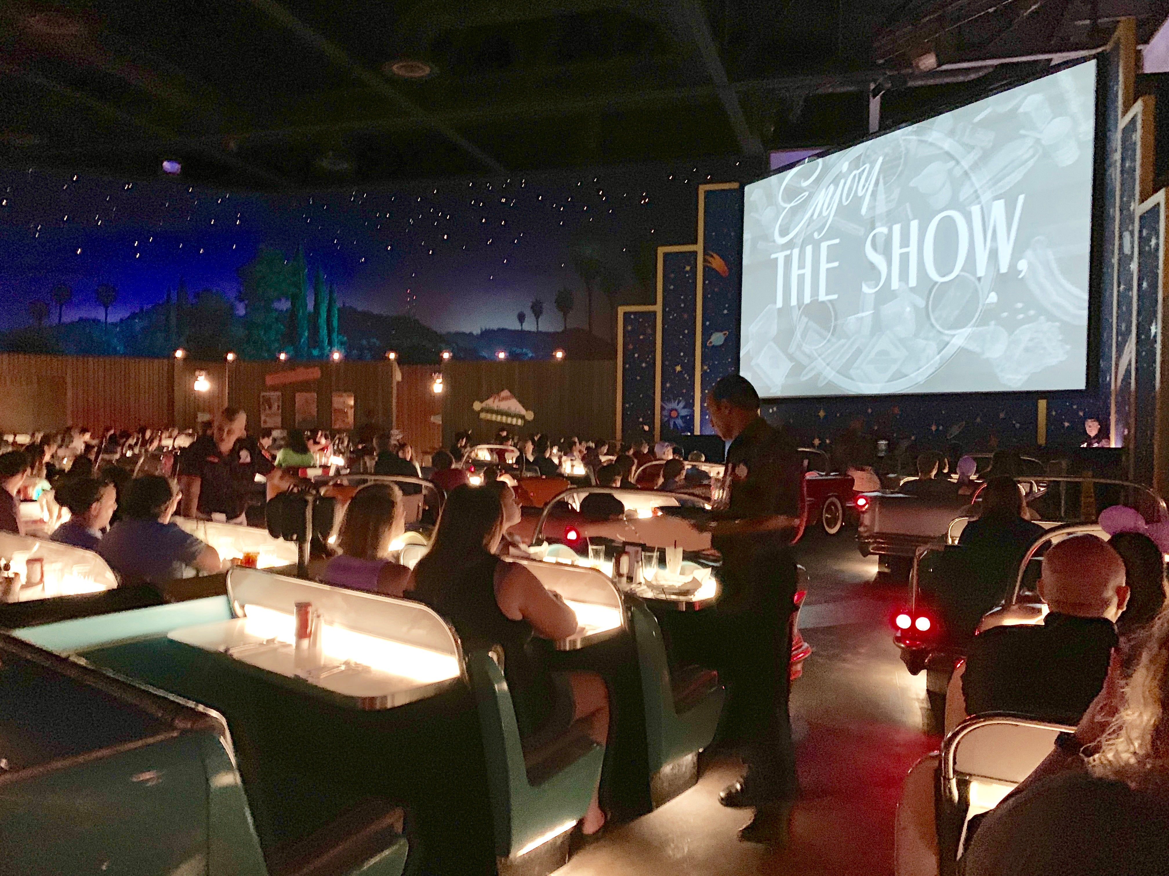 Diners sit at tables that look like seats in a car while a black-and-white movie plays on a screen in the background at Disney World's Sci-Fi Dine-In Theater Restaurant