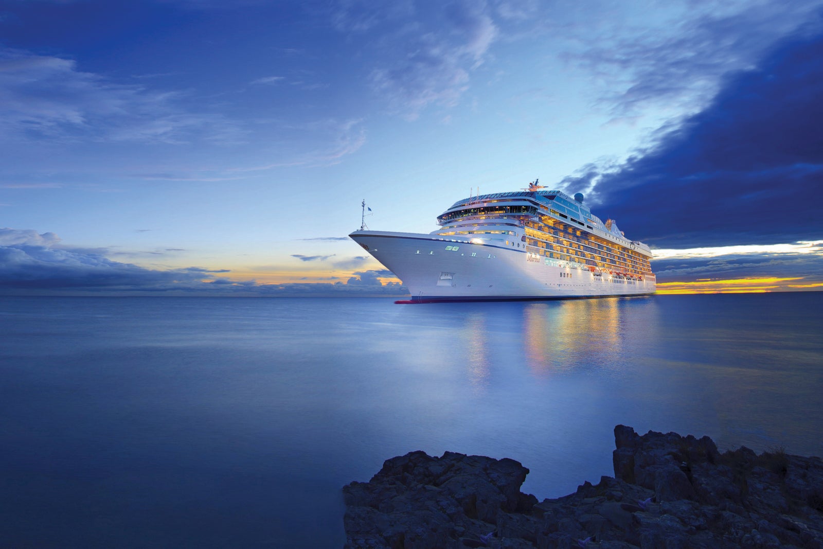 A cruise ship floats on the water near dusk with a nearly dark sky behind it and its interior lights lit