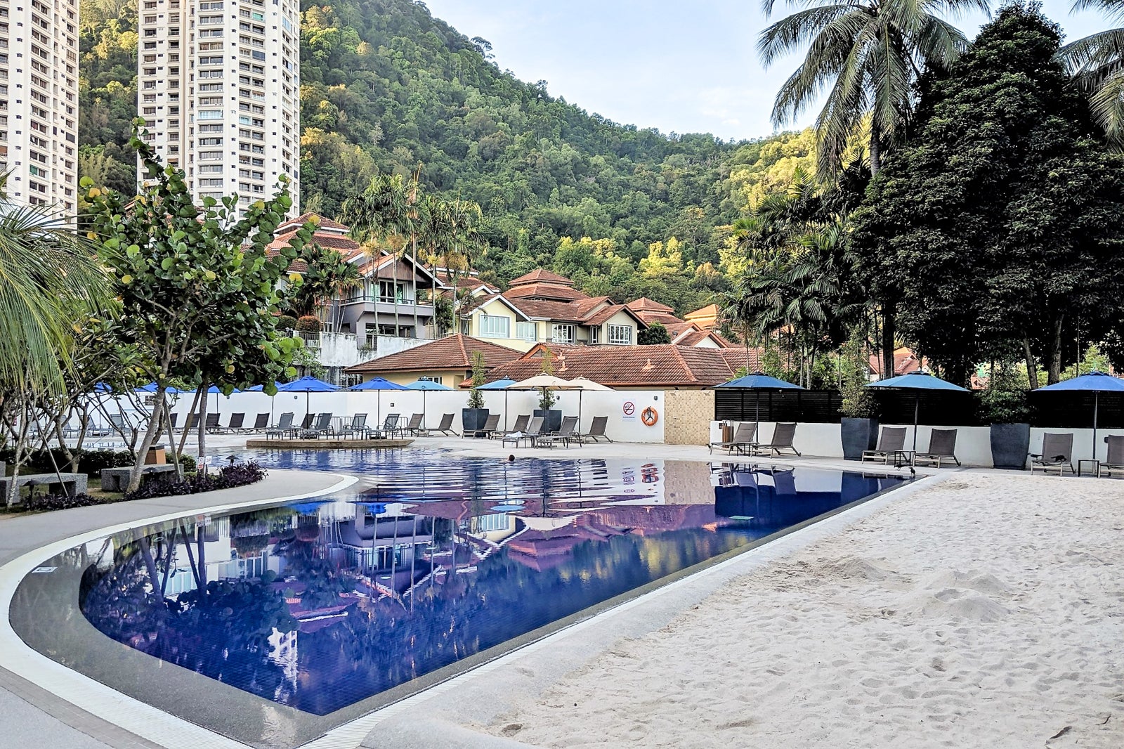 a calm hotel pool shows a reflection of homes, apartment towers and tree-covered hillsides beyond