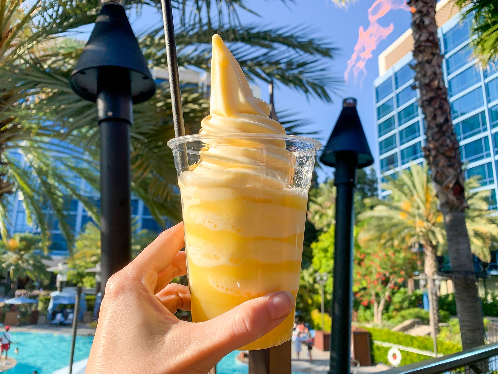 a hand holding a bright yellow Dole Whip in a clear cup in front of a pool at Disney World