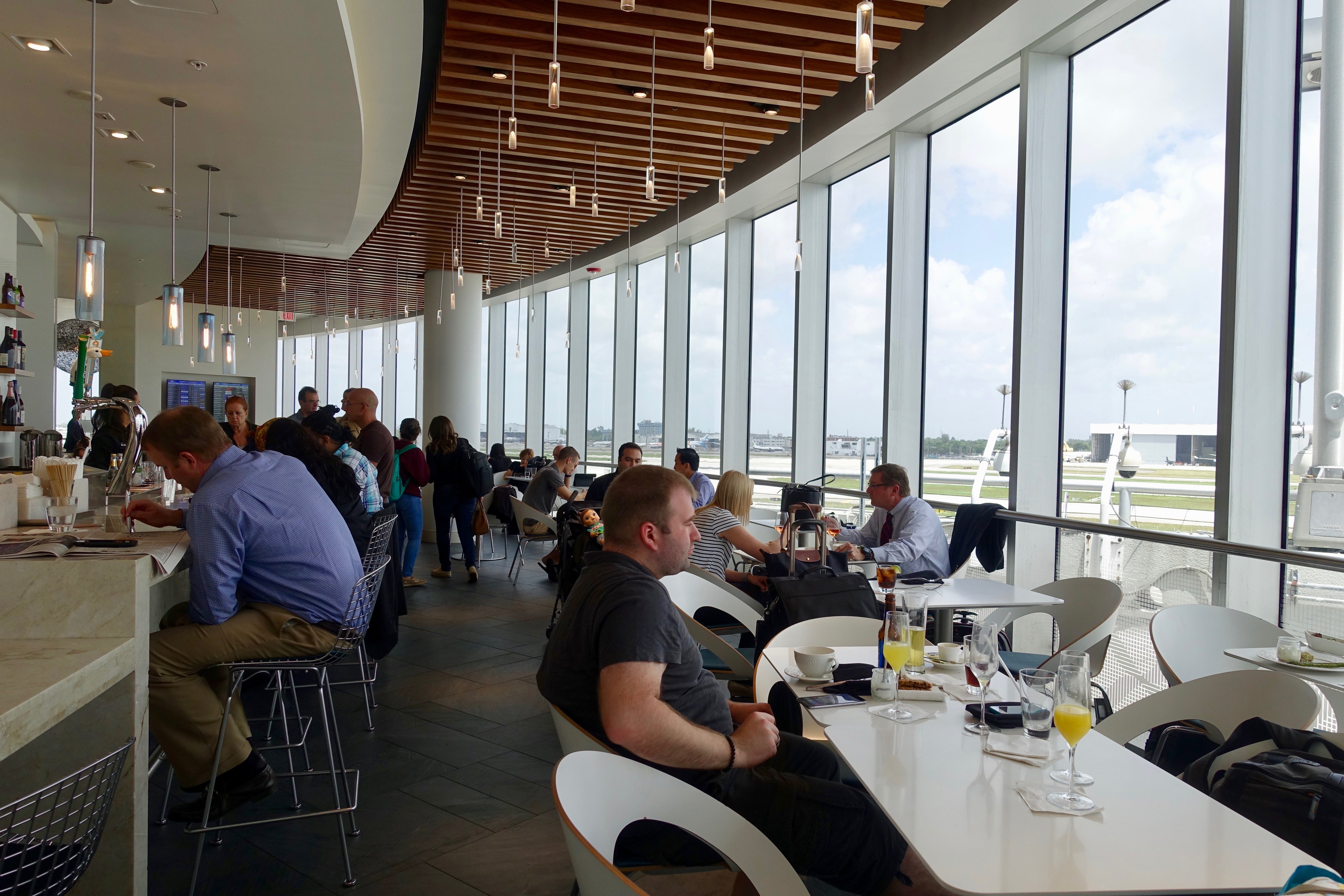 People sit at tables near viewing windows overlooking the runway at an airport lounge
