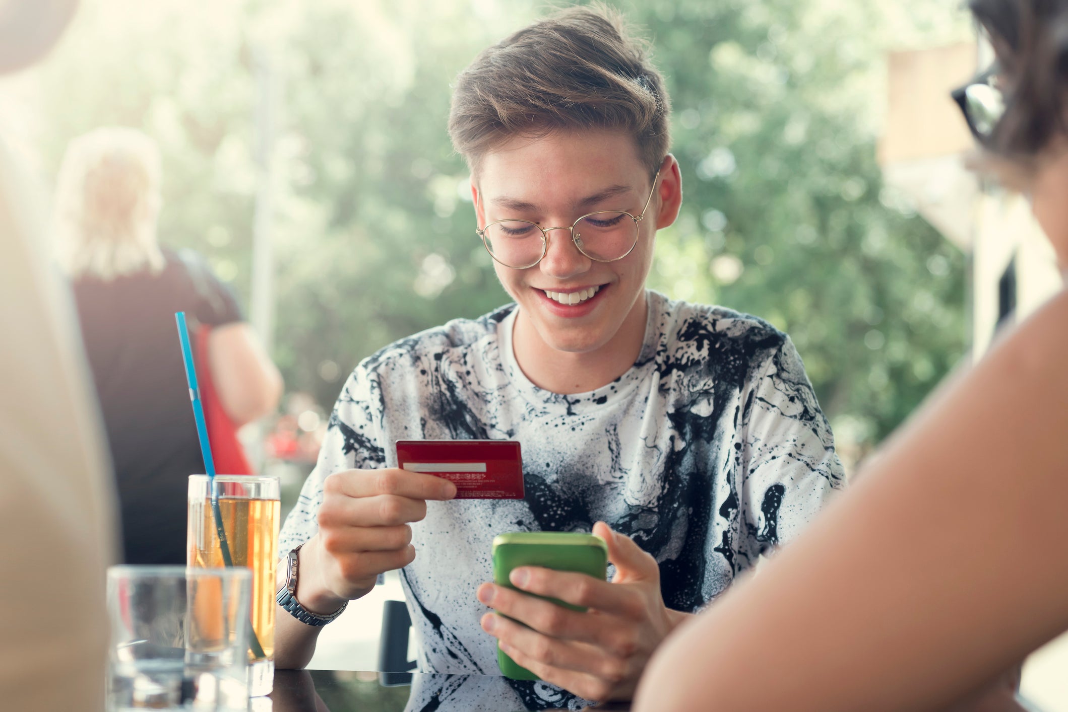 A teenager with short hair and glasses smiles while holding a credit card and mobile phone at a table at a restaurant