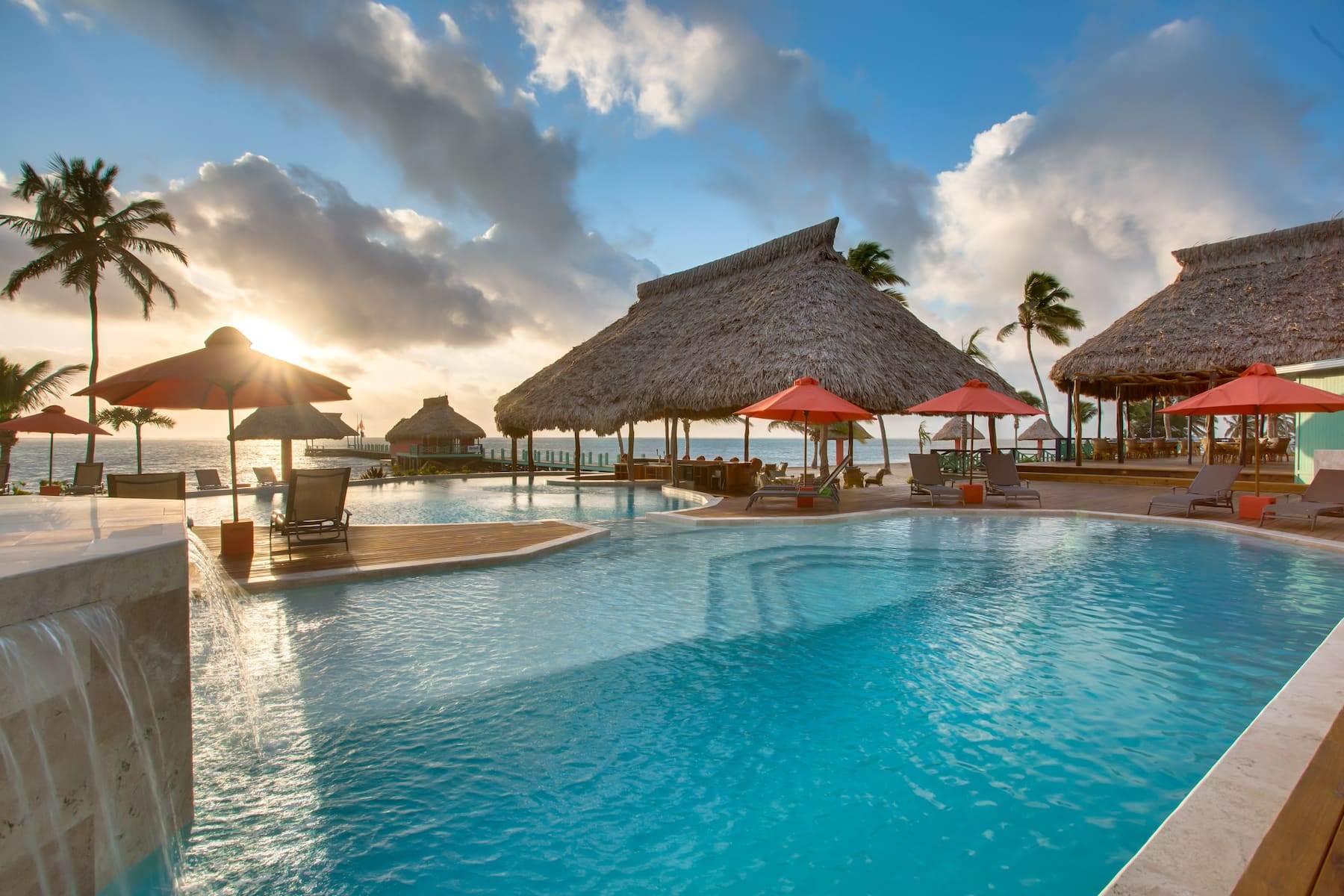 grass-thatched shelters sit between a hotel pool and the ocean; the sun sets in the background