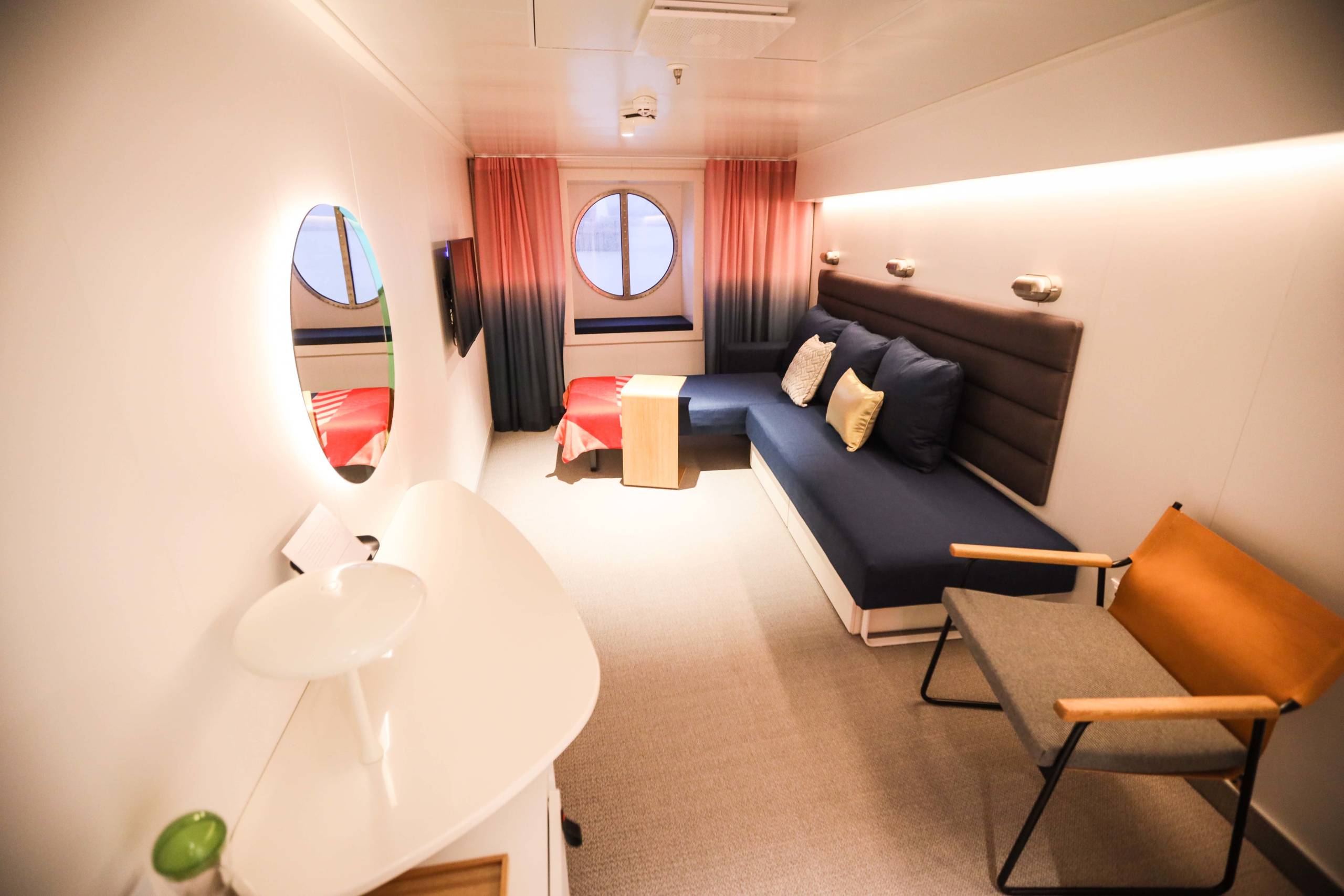 A cruise ship cabin with a white vanity, chair and blue L-shaped sofa