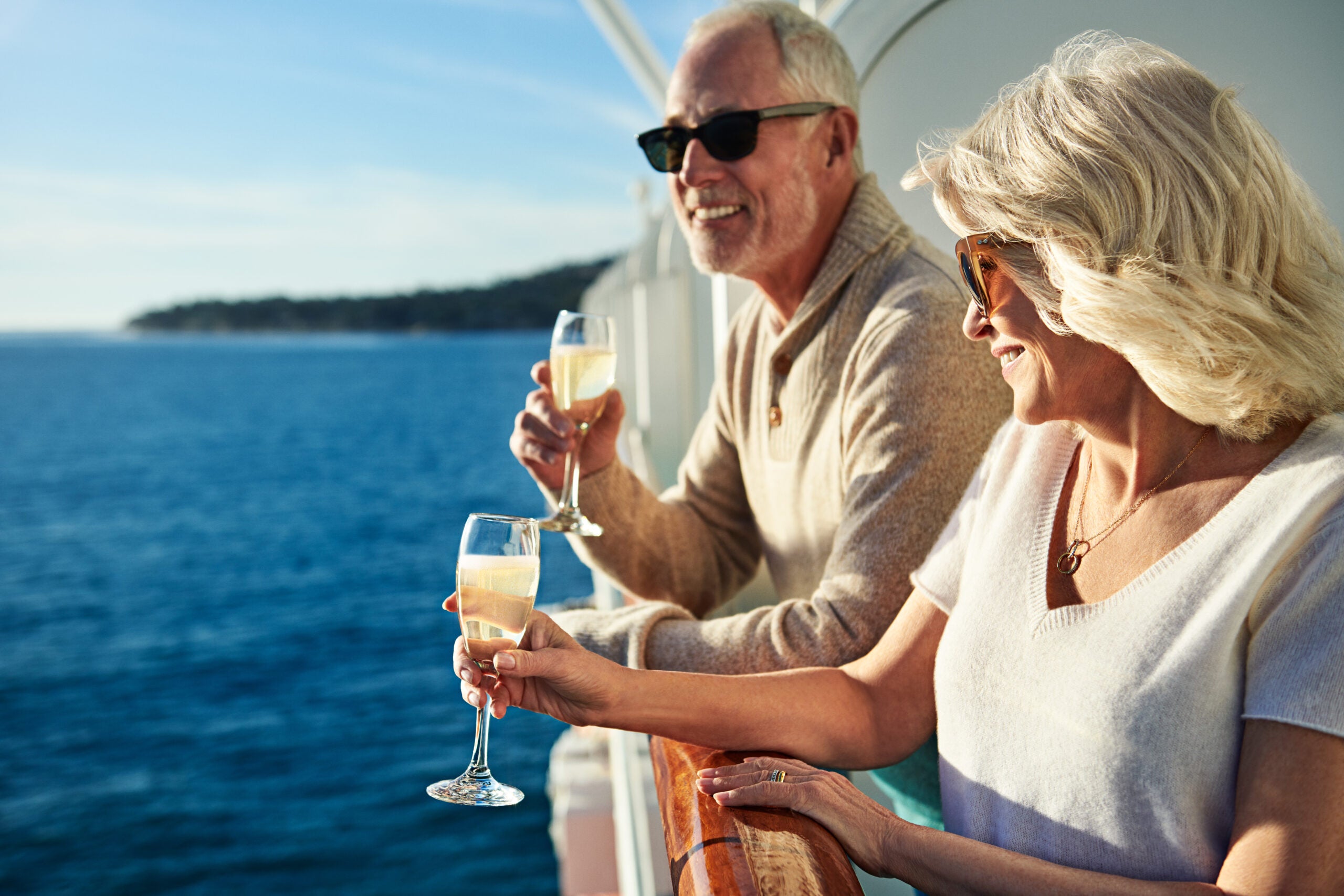 A smiling couple stands on a cruise ship balcony holding glasses of Champagne and watching the ocean glide by