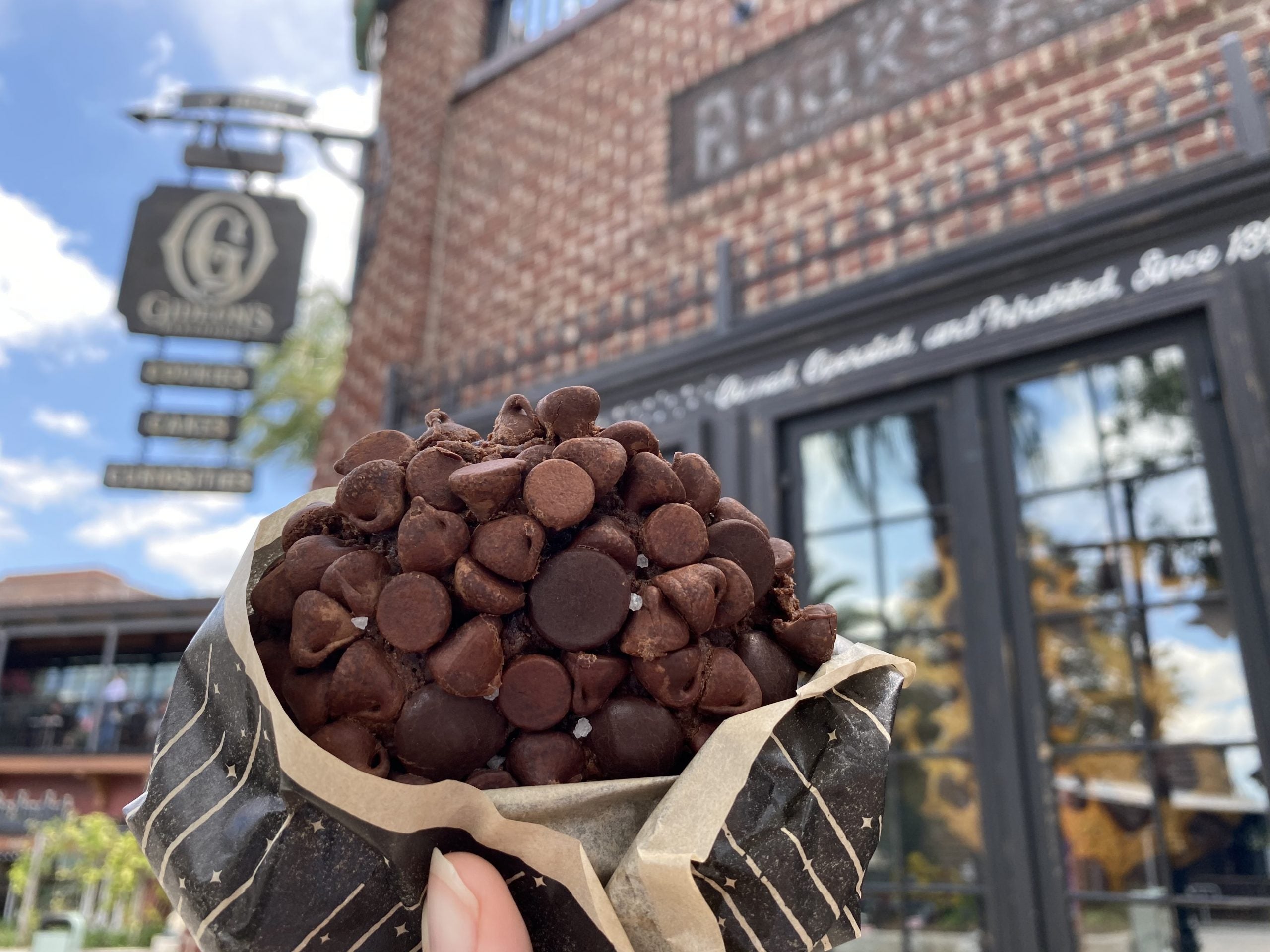 A hand holds a triple chocolate chip cookie in a beige wrapper in front of Gideon's Bakehouse in Disney Springs at Walt Disney World