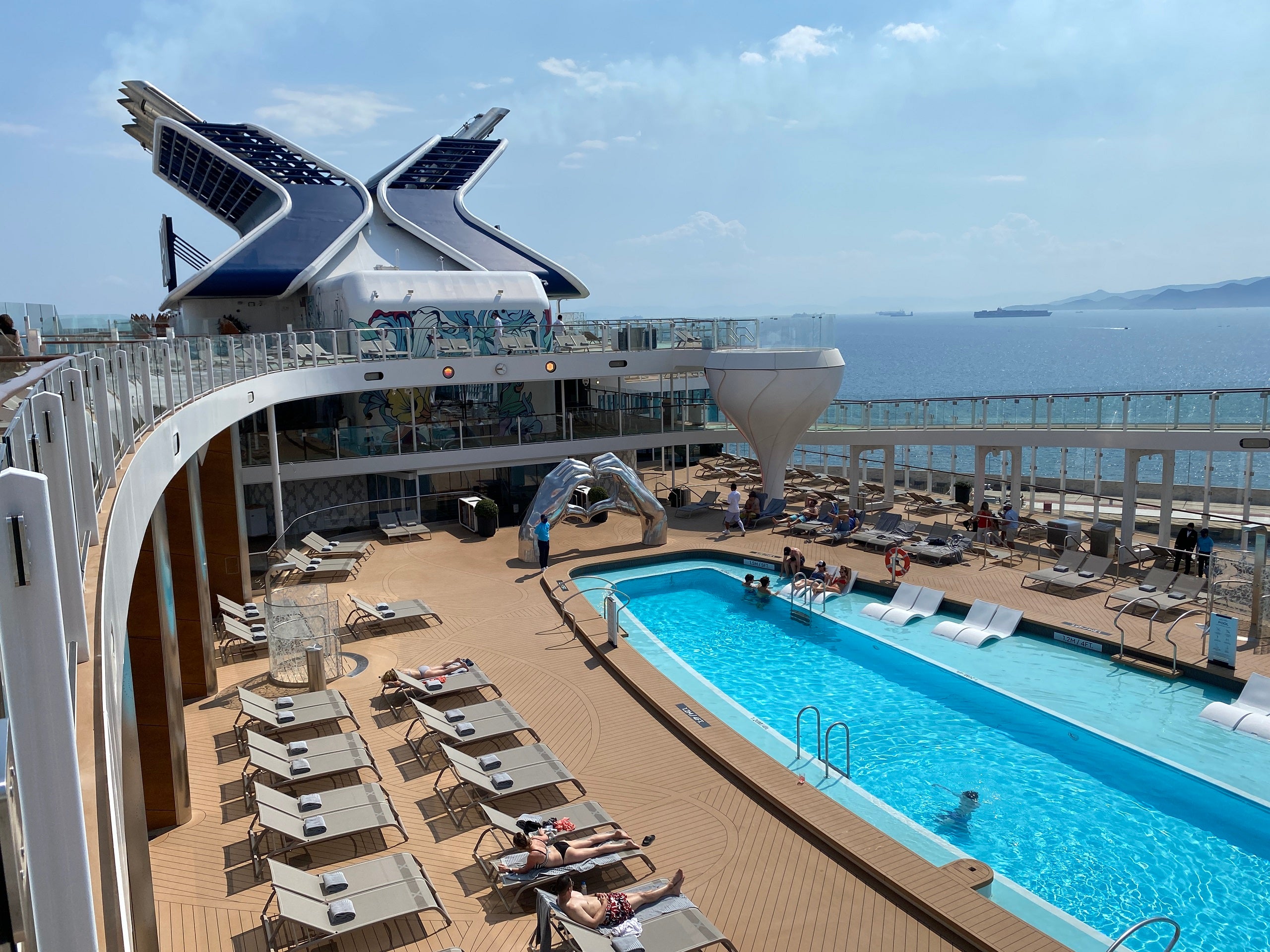 An aerial view of a cruise ship pool deck featuring a blue pool filled with water, a wooden deck with deck chairs, a blue funnel and a silver sculpture of two hands forming a heart