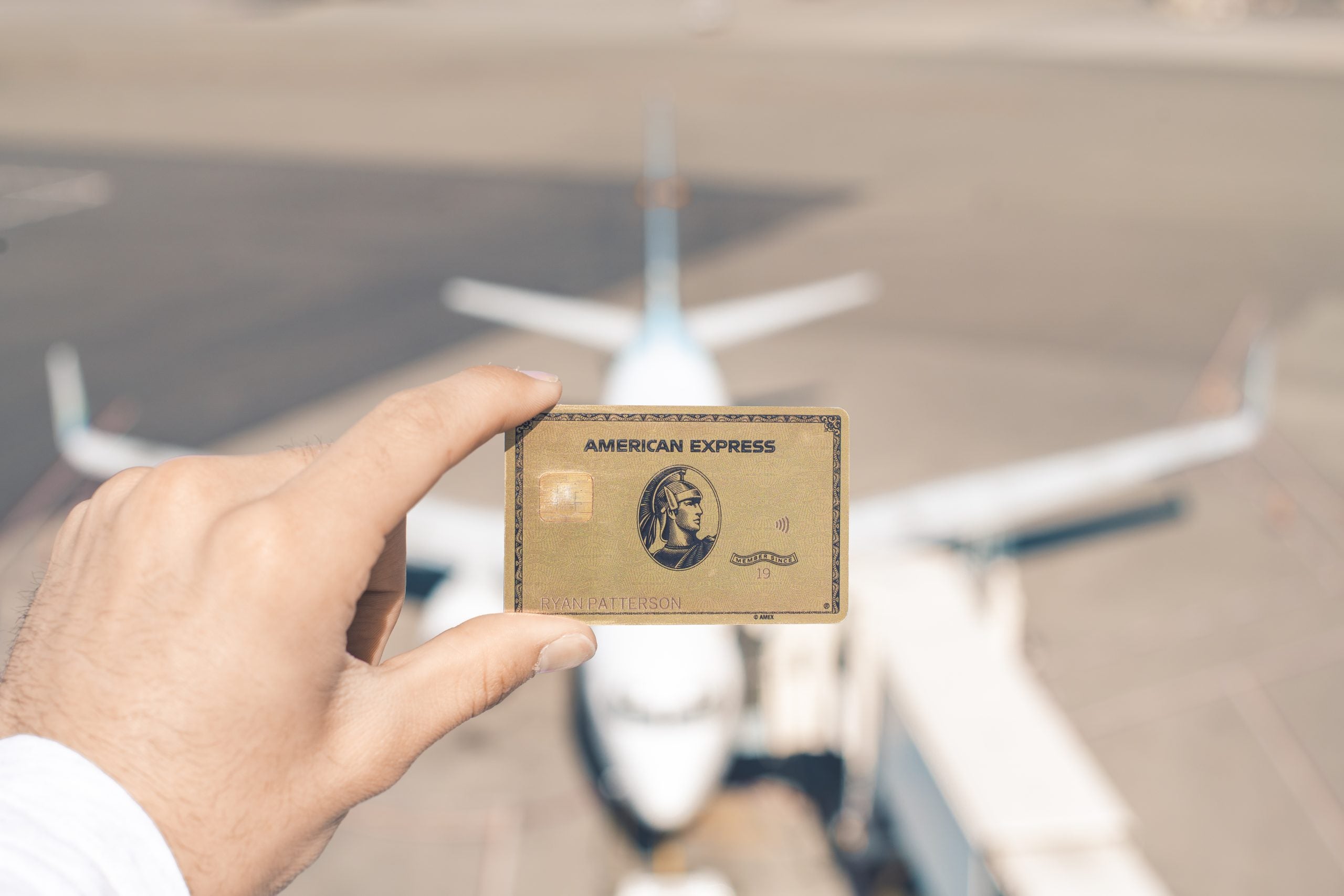 a hand holds the Amex Gold Card with a plane visible in the background