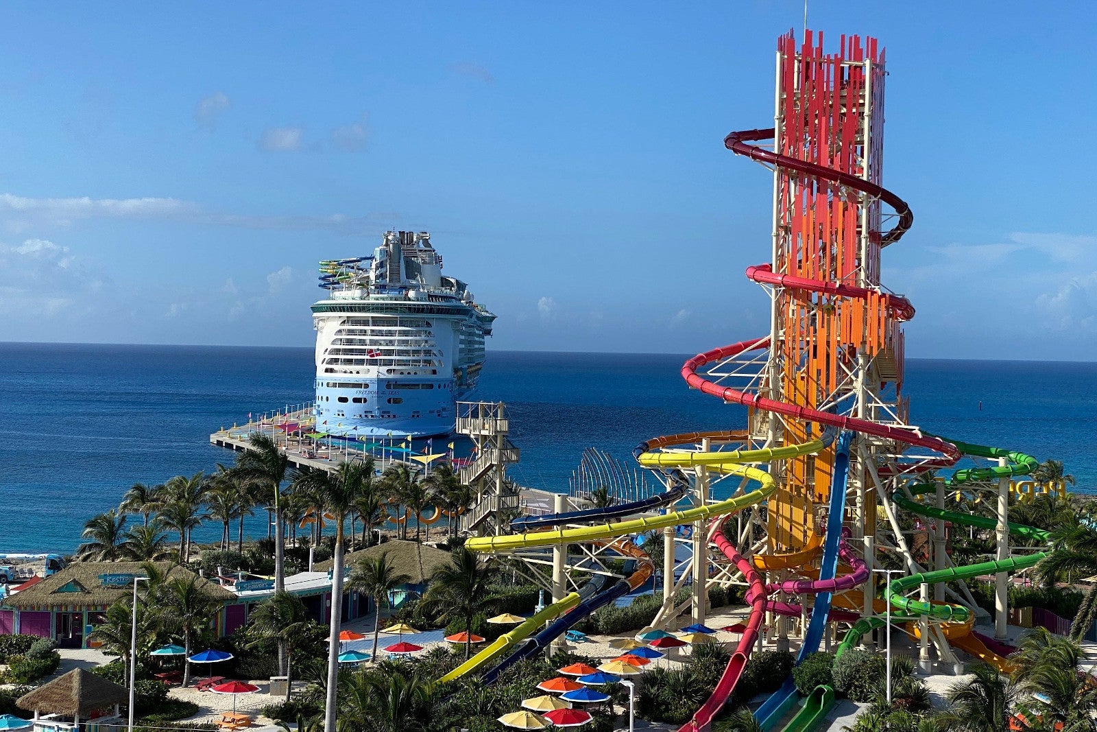 An island with beach umbrellas, waterslides, palm trees and a cruise ship docked in the background