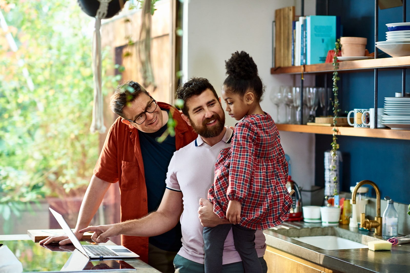 a couple holds a child in their kitchen while one of the parents uses a laptop