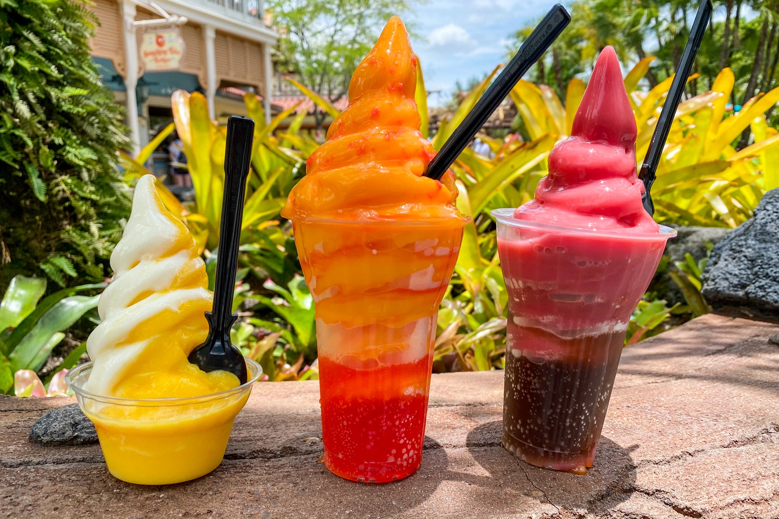 Three clear cups lined up on a rock wall are filled with yellow, orange and red Dole Whip flavors