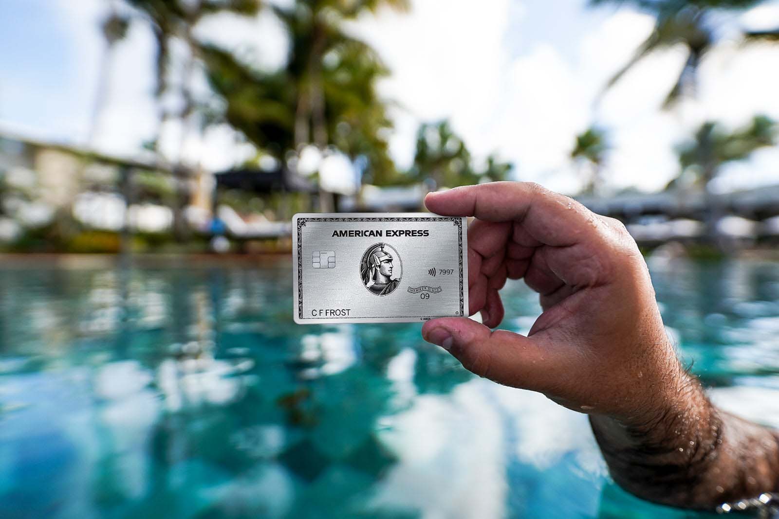 A hand holds a credit card with a pool and palm trees in the background