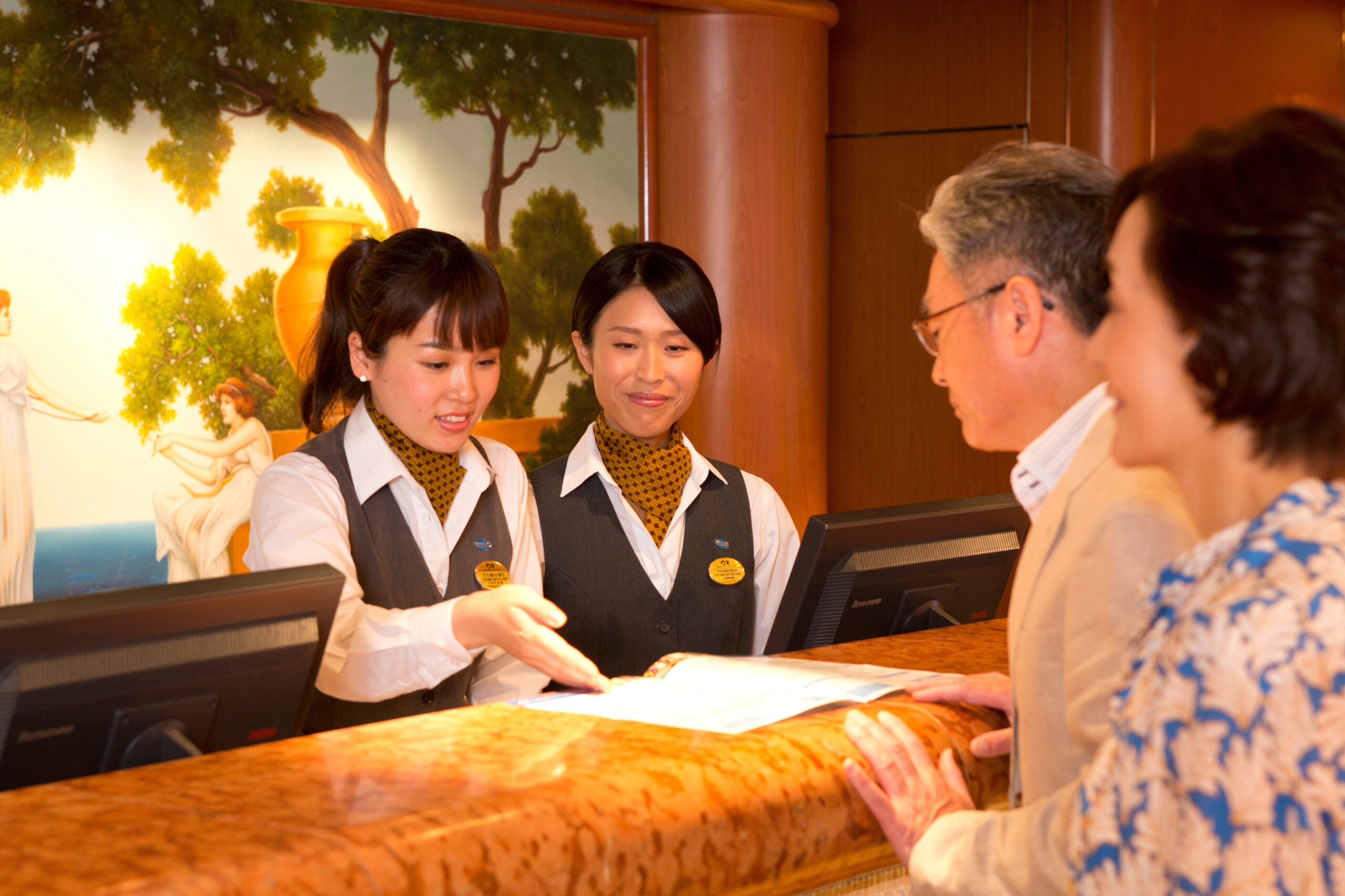 Two crew members behind a guest services counter assist two cruise passengers with paperwork