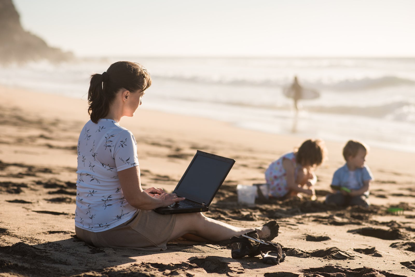 woman sitting on beach using laptop while kids play in the sand