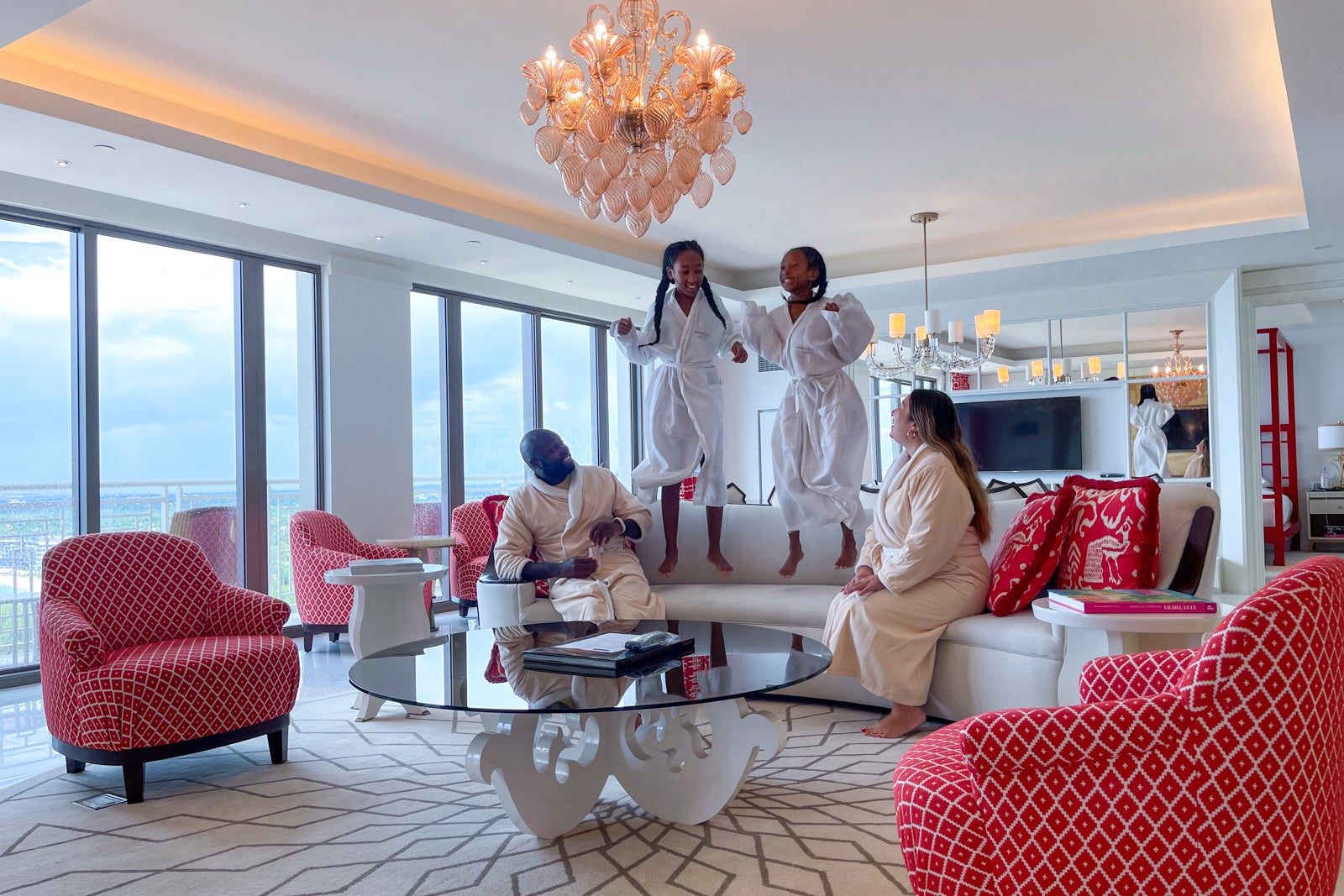 2 children jump on a sofa in a hotel room while their parents watch; all 4 people are wearing bath robes
