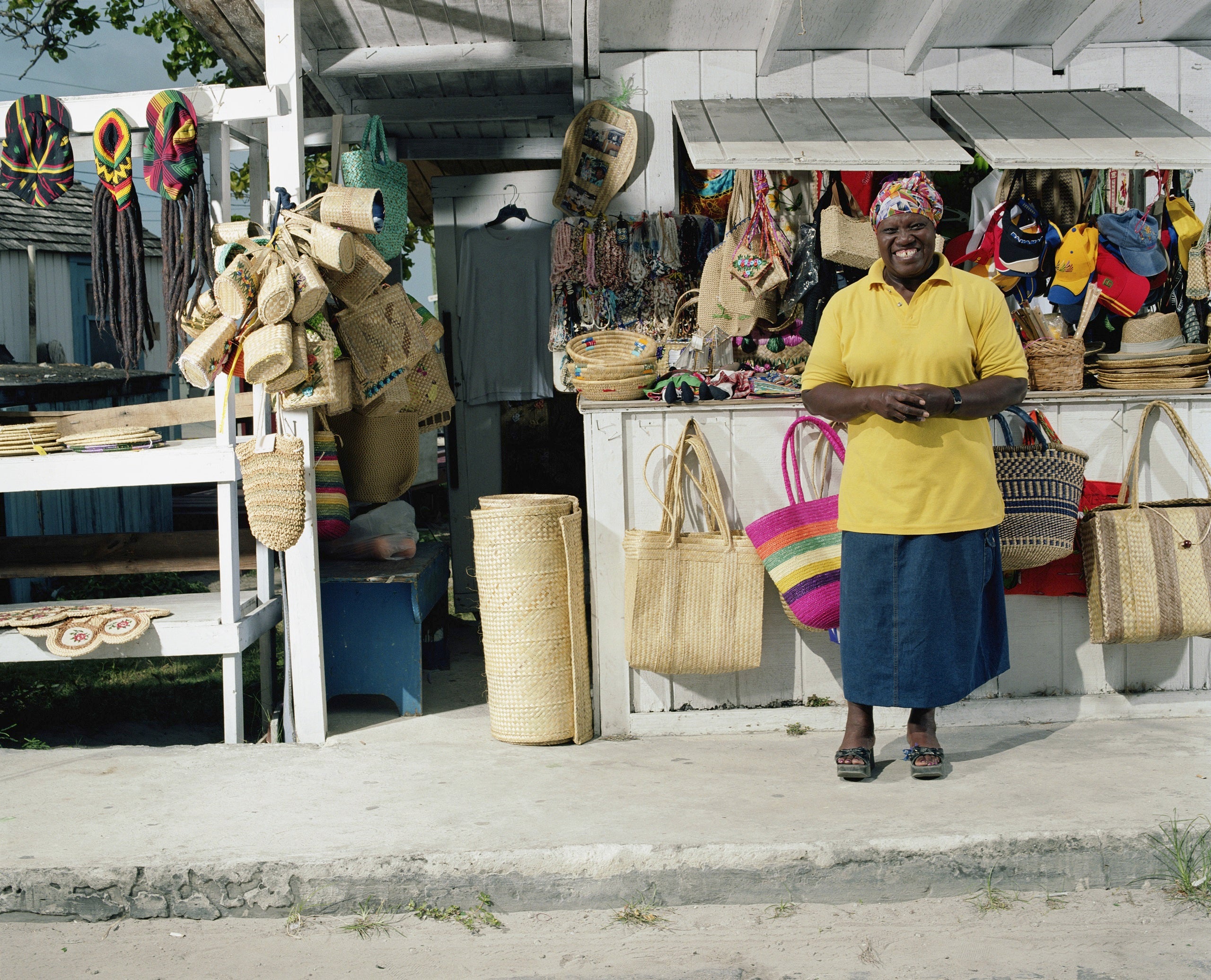 A smiling Bahamian woman in a yellow shirt standing in front of a stall at the straw market in Nassau