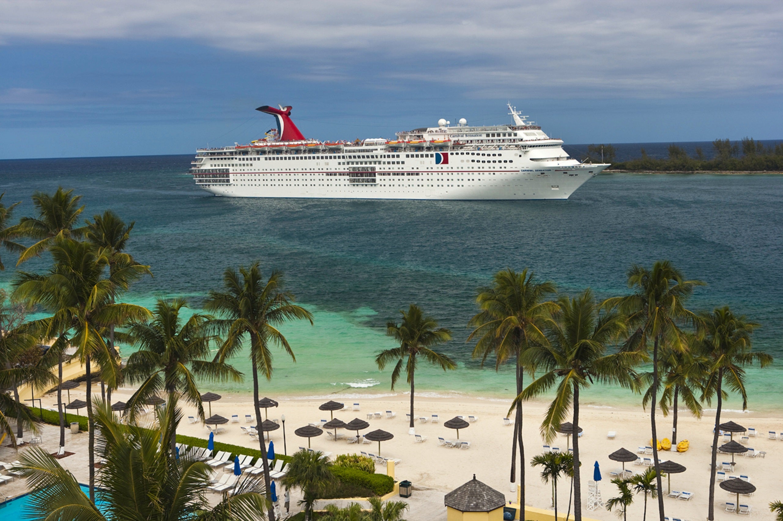 Aerial shot of a Carnival cruise ship anchored in the water near a beach with white sand, teal water and palm trees