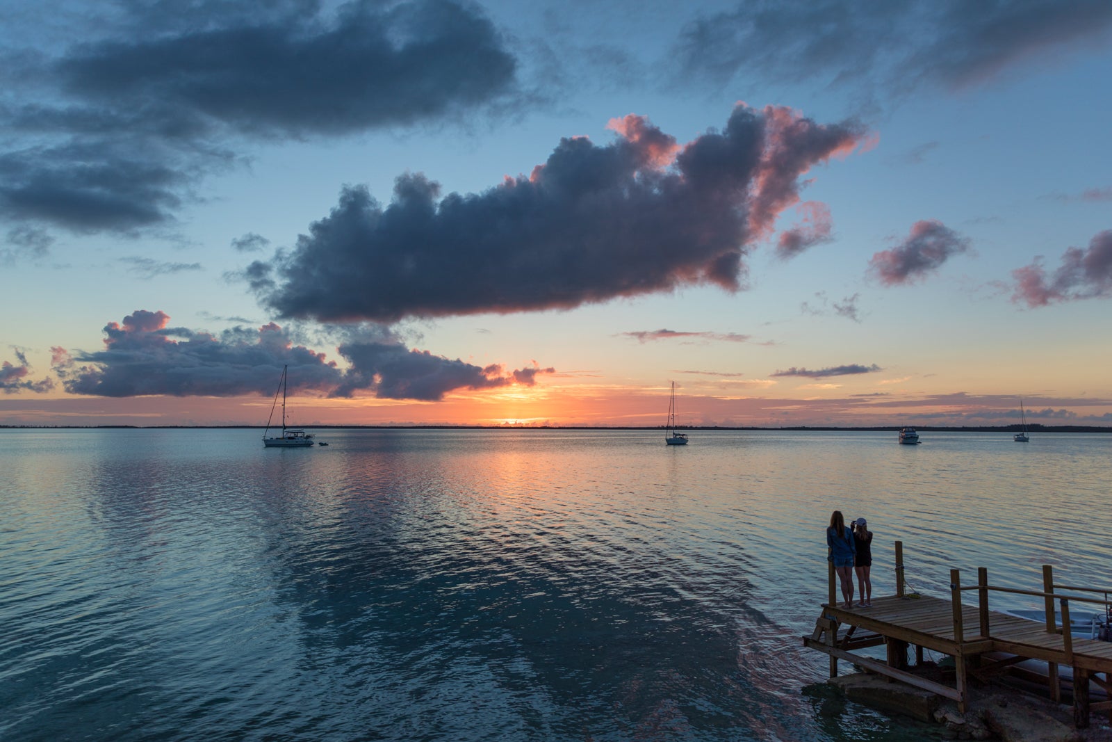 A silhouette of two people sanding on a dock watching sailboats at sunset