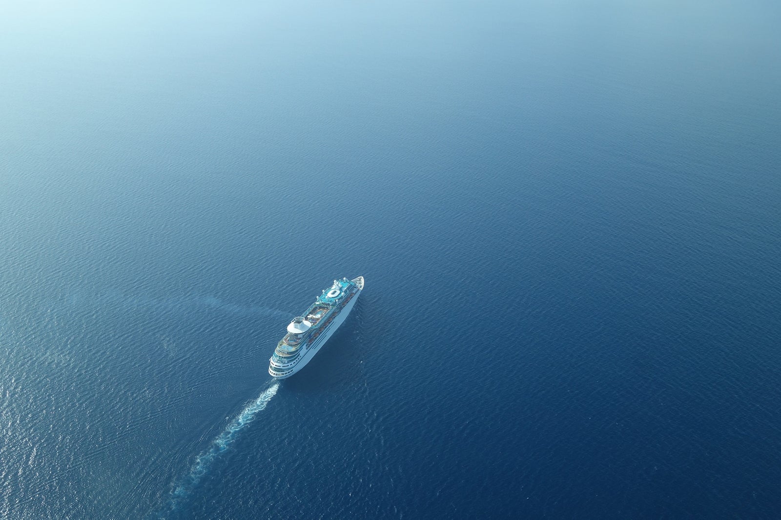 Aerial view from high above of a white cruise ship sailing on blue water with a white wake behind it