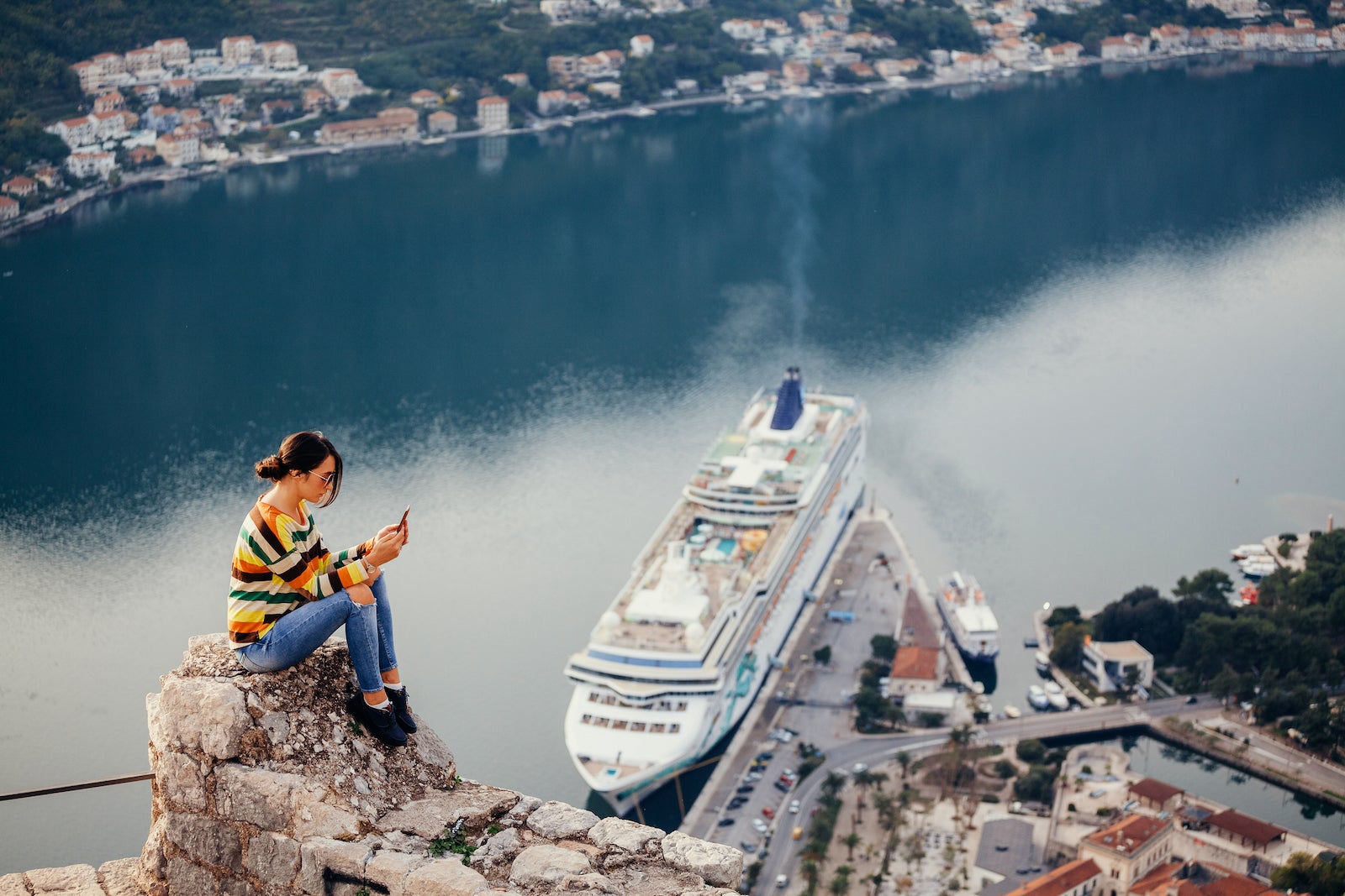 Woman sitting on a hill with a cell phone overlooking a cruise ship in the water