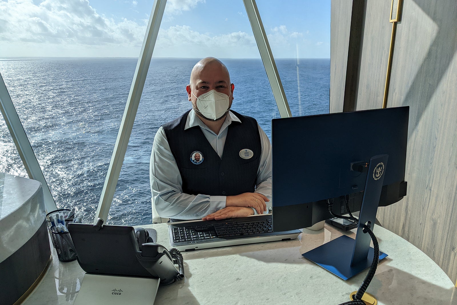 Bald man in vest sitting at desk with sea behind him