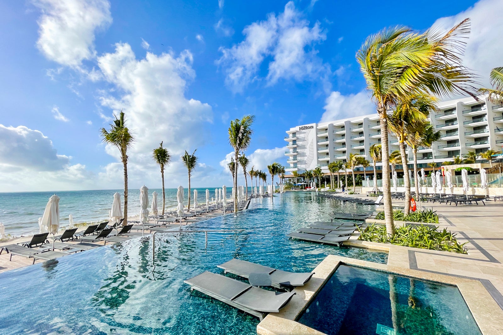 photo of lounge chairs and palm trees near a pool, with a hotel and ocean in the background