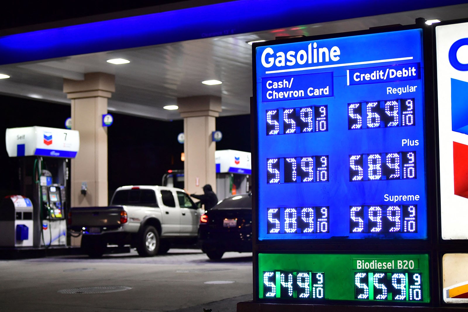 photo shows people and cars at a gas station, with a sign displaying prices in the foreground--including a discount if paying cash