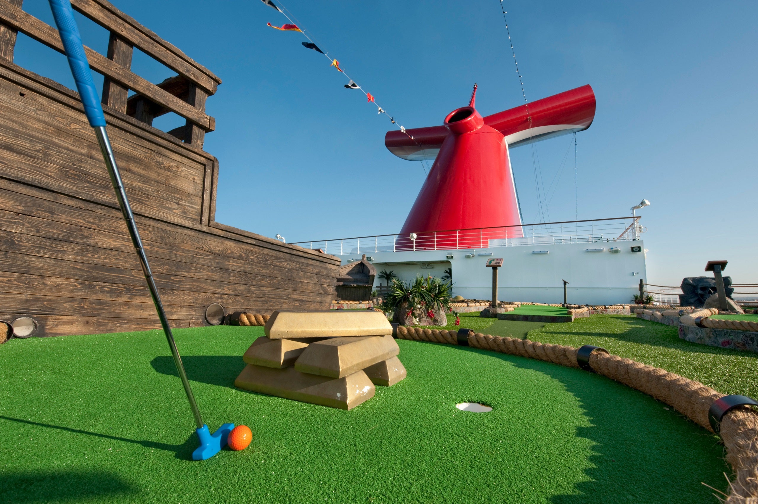 A blue club and red ball rest on a mini-golf course with a Carnival cruise ship red funnel in the background