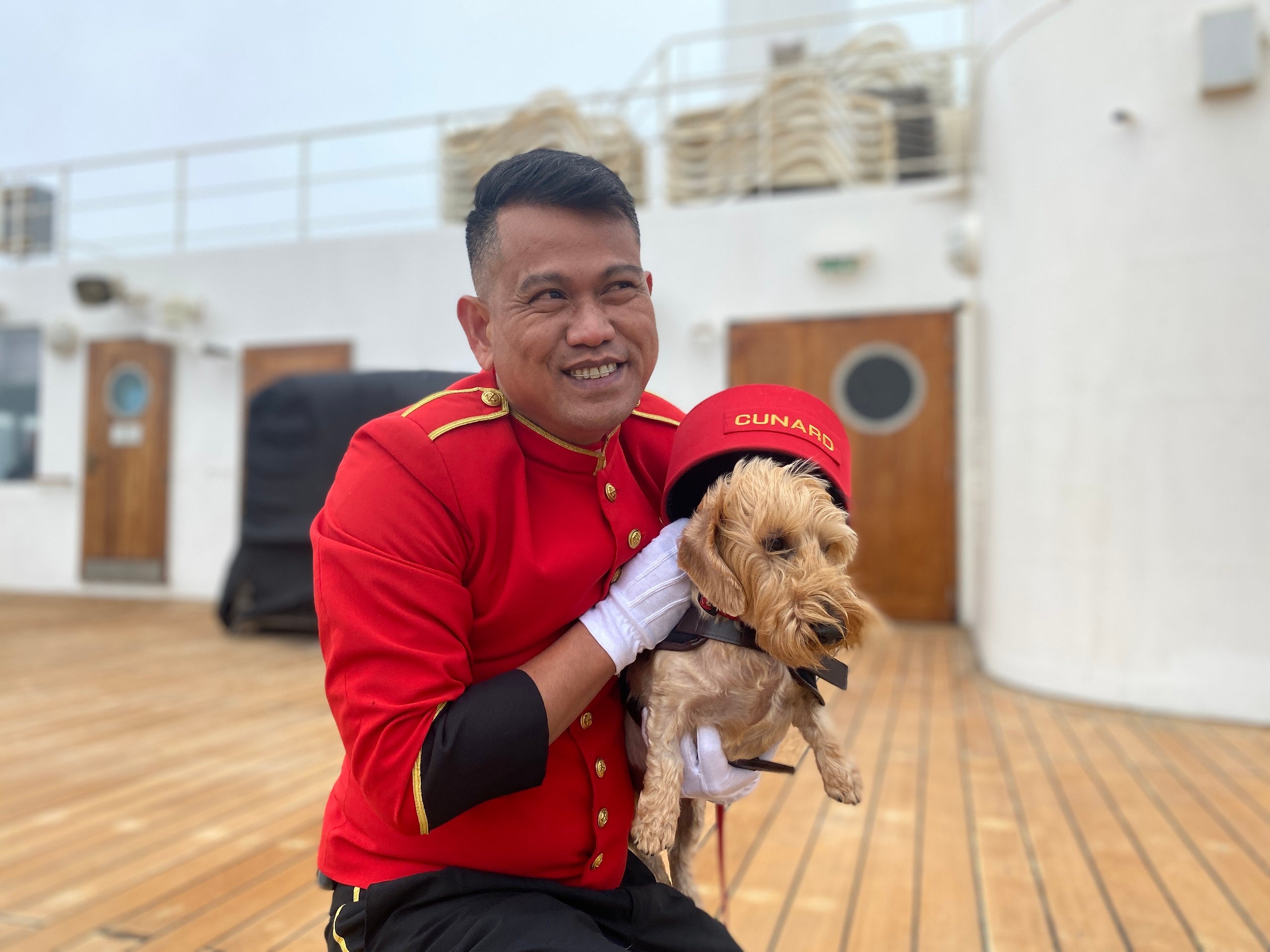 Queen Mary 2's kennel master holding a dog in a red bellhop hat on the deck of the ship