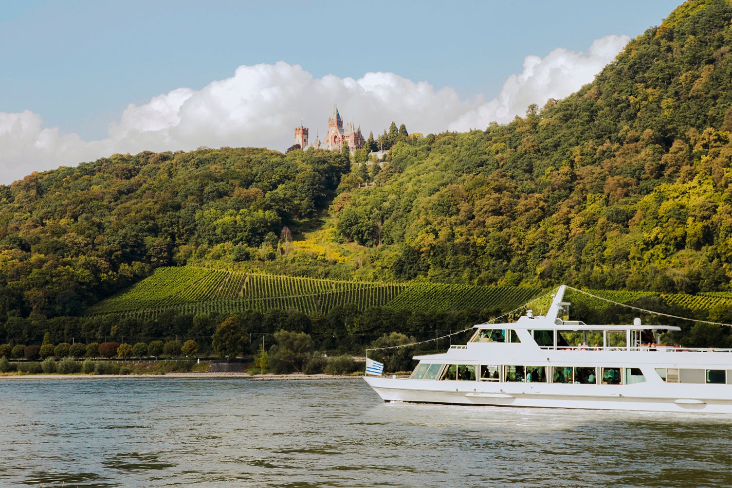 A river cruise boat sailing into the frame of the photo along the Rhine with green hills in the background