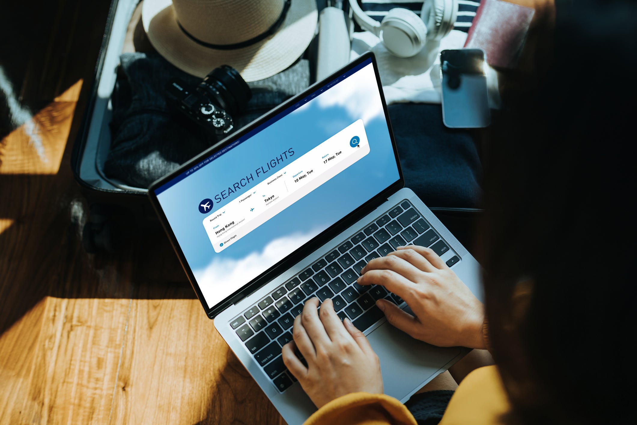 Young Asian woman shopping online for flight ticket on airline website with laptop at home, sitting next to suitcase with sun hat, camera, clothings, headphones, smartphone and passport. Getting ready for vacation. Travel and vacation concept