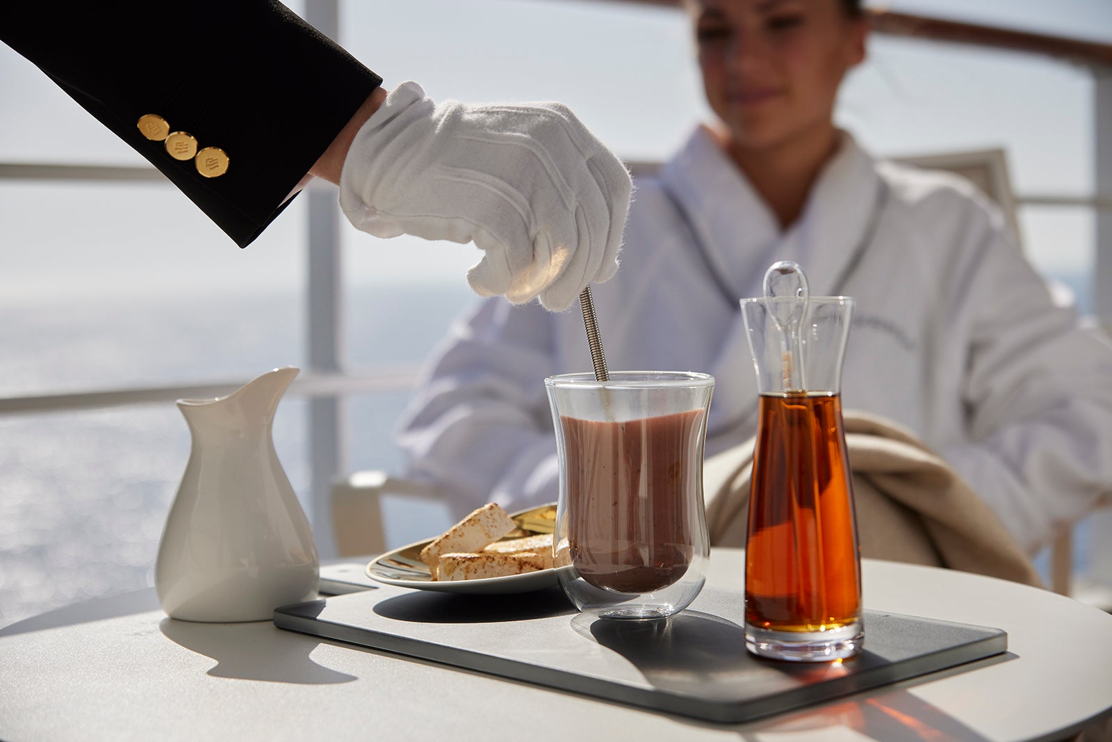 The white-gloved hand of a butler stirs tea on a cruise ship balcony as a passenger in a bathrobe looks on in the background