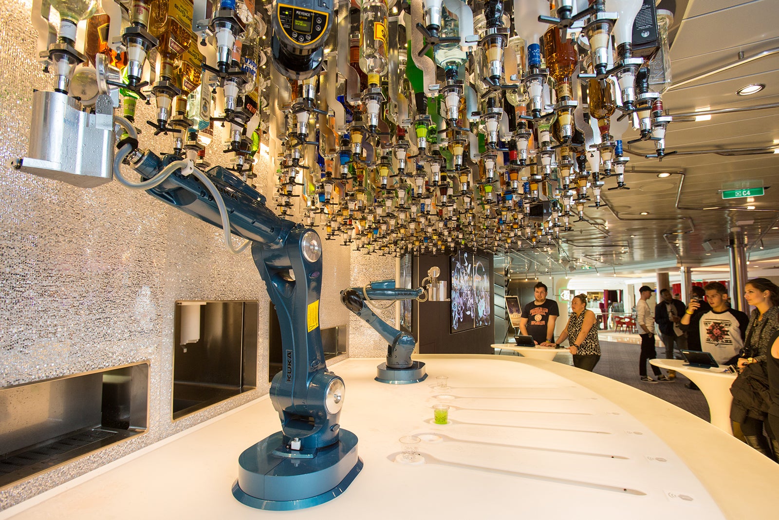 Two robotic arms on top of a bar with dozens of alcohol bottle suspended upside down overhead