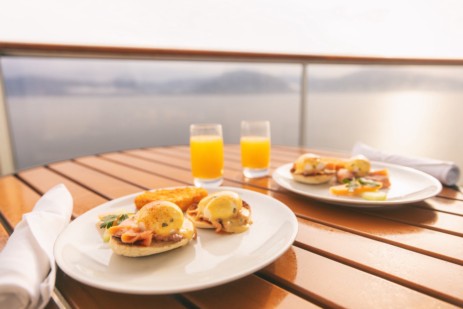 A slatted table on a cruise ship balcony holds two plates of breakfast food and two glasses of orange juice.