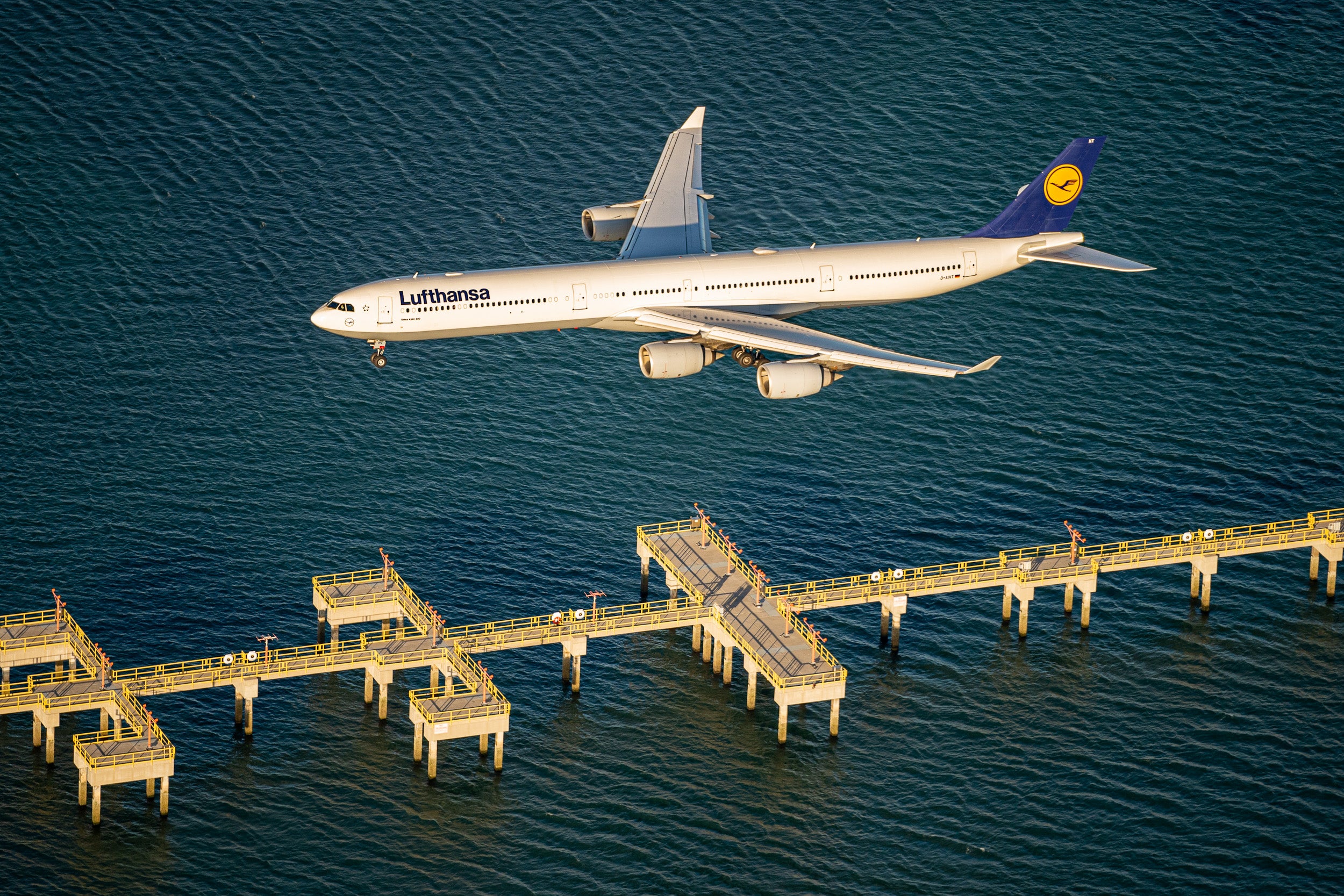 A Lufthansa A340-600 flies over landing marker beacons on a pier on the water just off of the edge of Boston Logan airport.