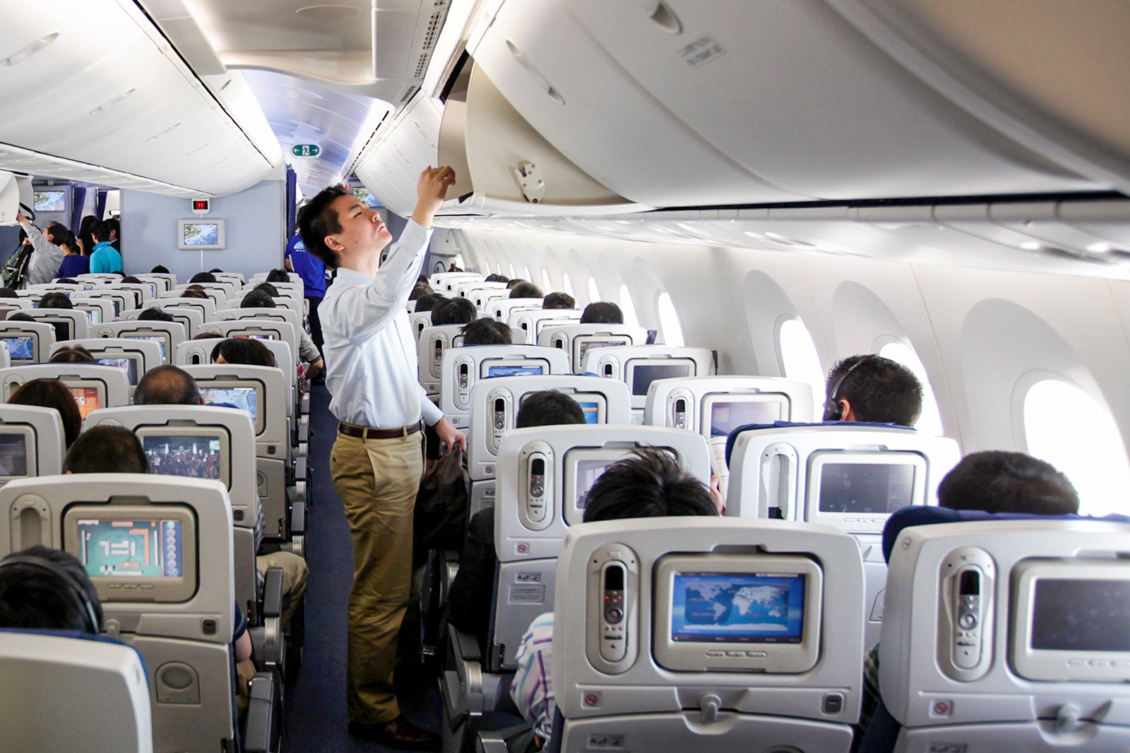A person standing up in the aisle and looking in the overhead bin 