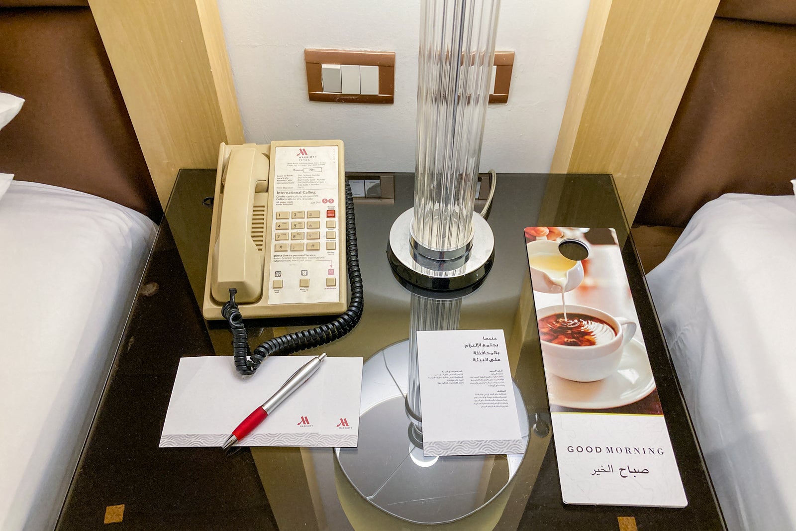 items on a night stand between two beds, showing an outdated phone and wall socket