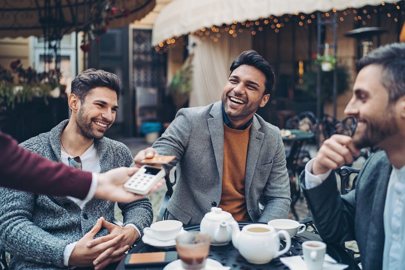a smiling group of friends sits at a table while paying with a credit card after dinner