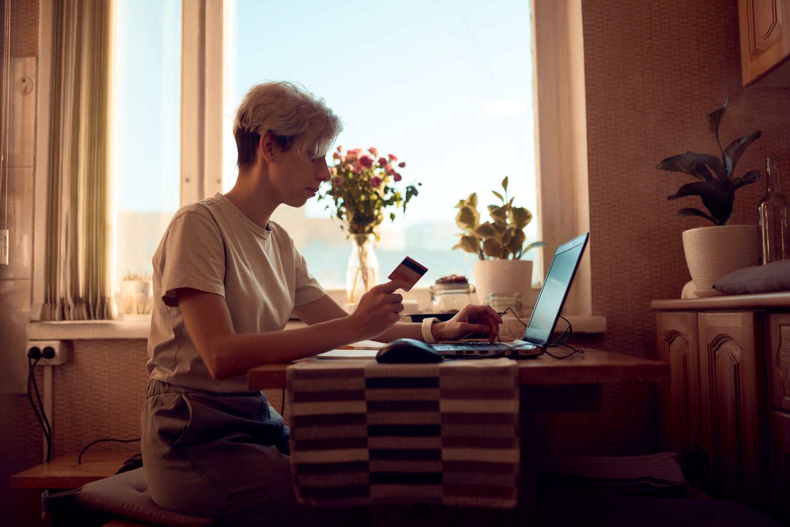 a person sits at a small table near a window, holding a credit card and reading information on a laptop