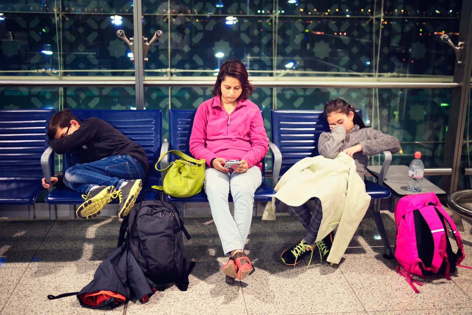 A family waits in an airport; the children are trying to sleep while the mom plays on her phone