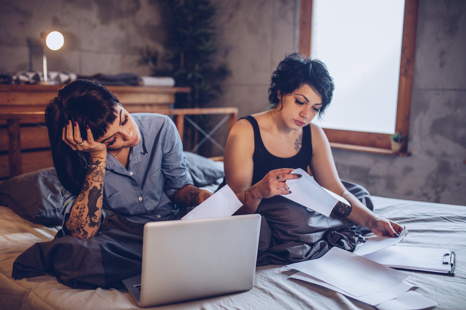 a couple sits on a bed looking at documents and a computer; they look frustrated