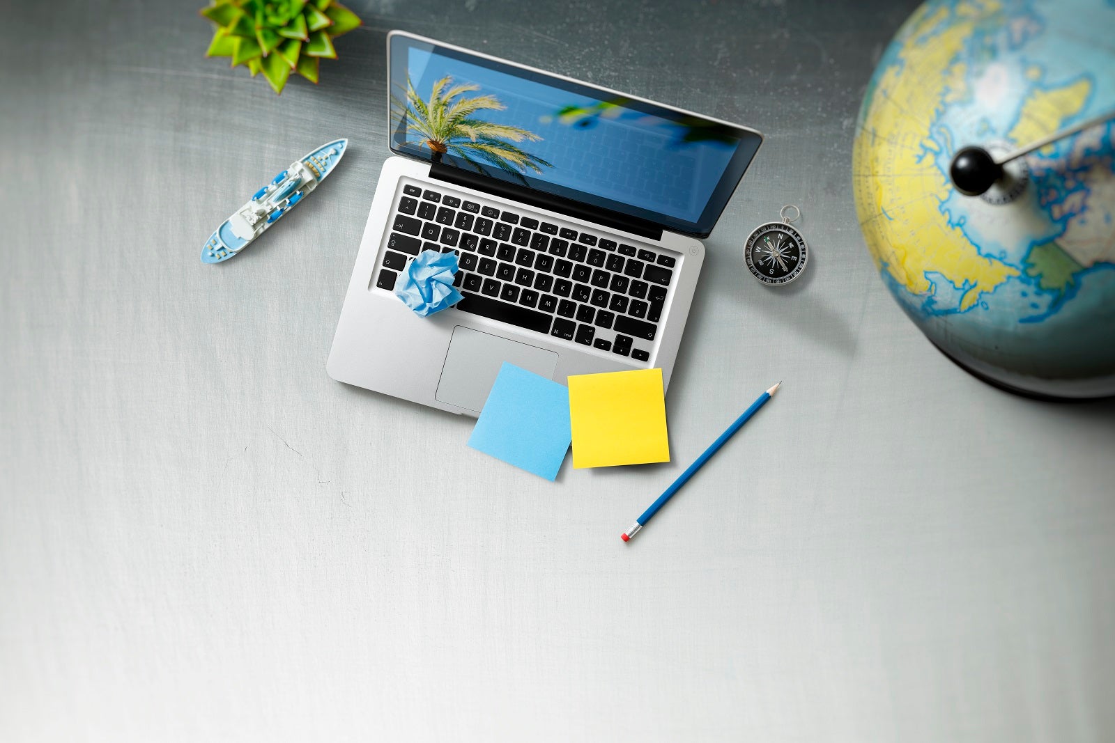 A view from above of a silver laptop on a table with a toy cruise ship on one side, a pencil on the other and sticky notes on top of it