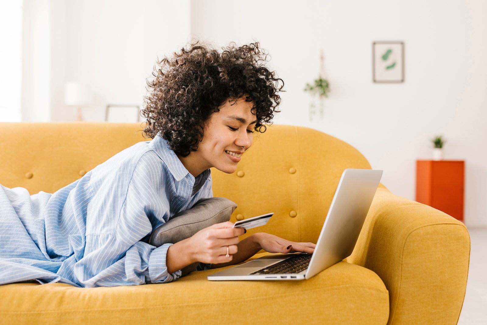 Smiling woman holding credit card using laptop lying on sofa in living room at home