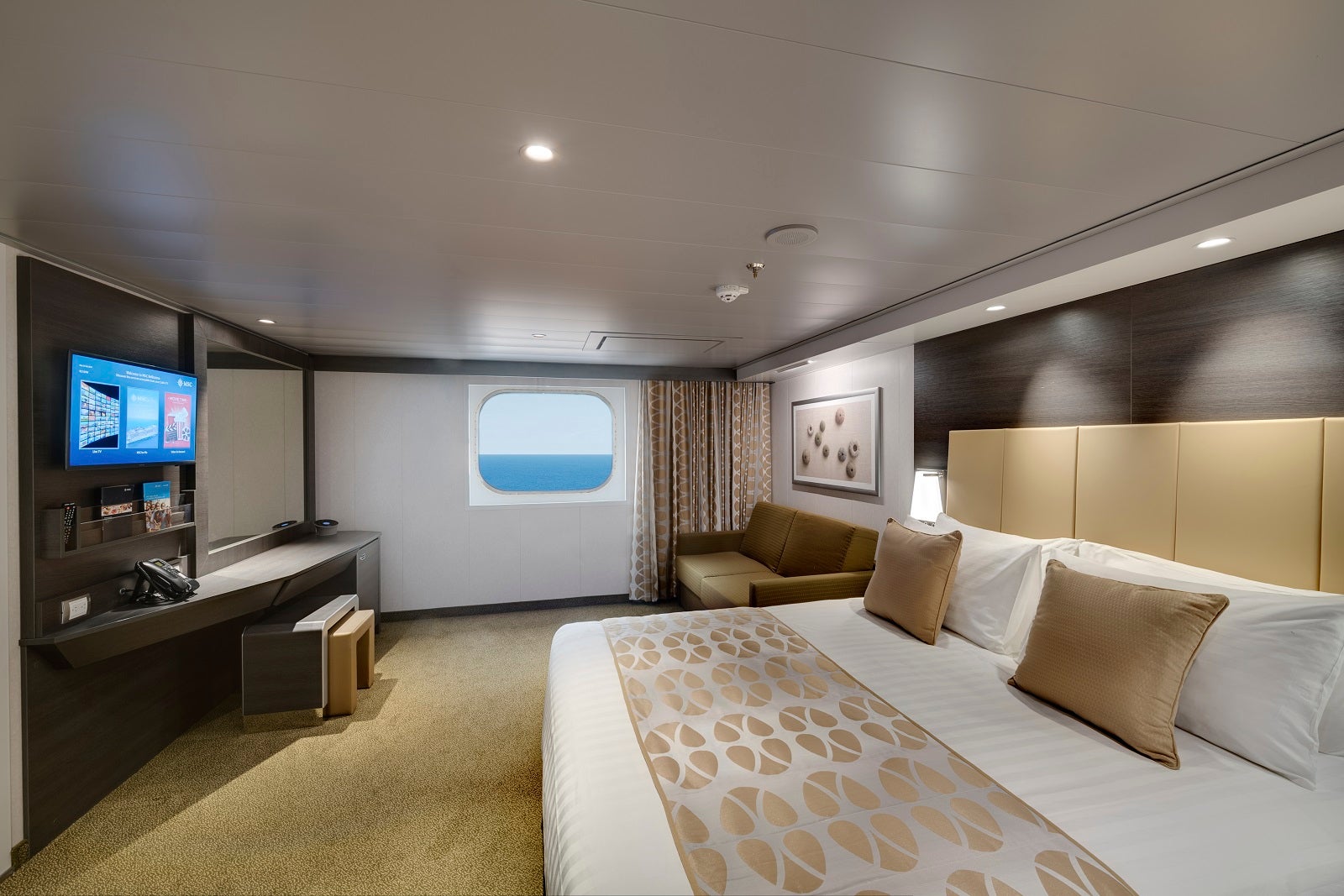 A cruise ship cabin with a bed, sofa, desk, TV and window