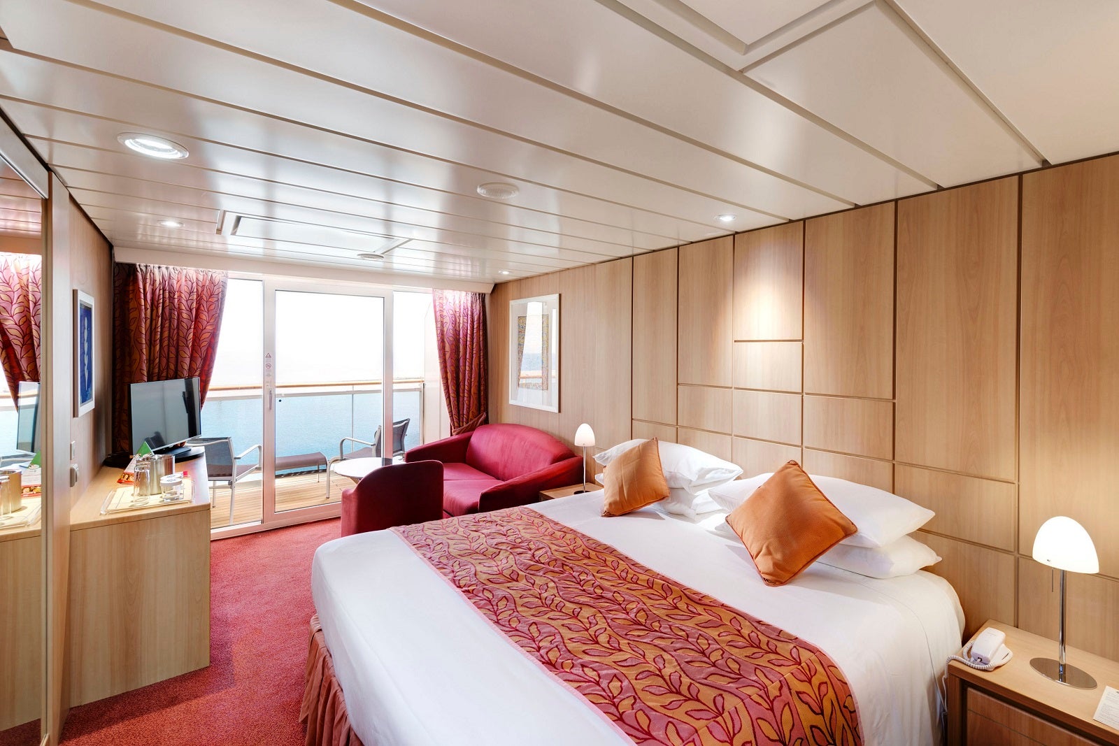 A cruise ship cabin with red decor, wood accents and view of the balcony