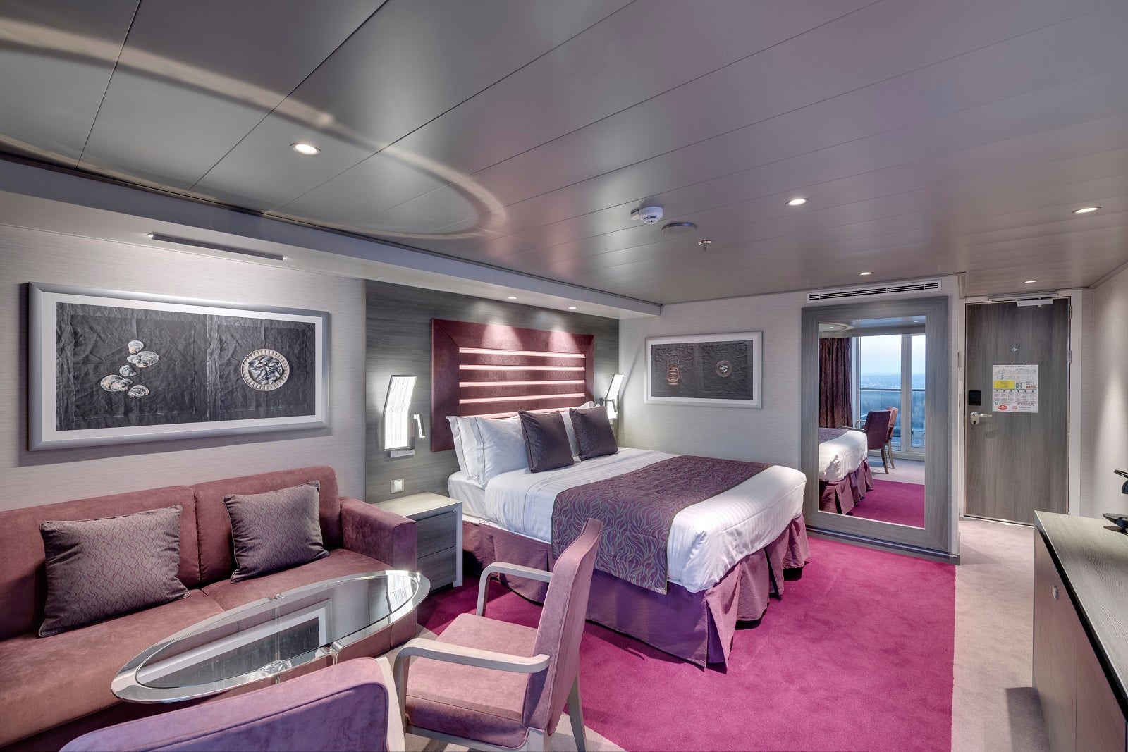 A cruise ship cabin decorated in mauve colors with a bed, sofa, table, chairs and a large mirror