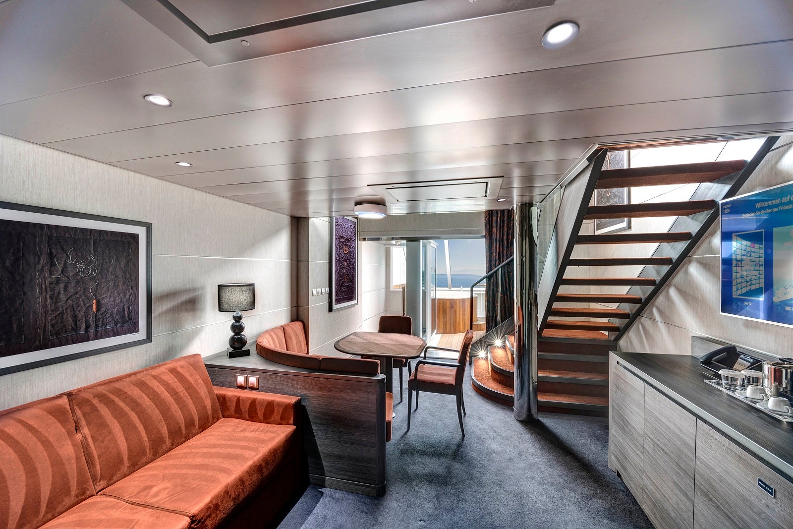 A cruise ship cabin with a sofa, table and chairs, and a set of stairs leading to another level
