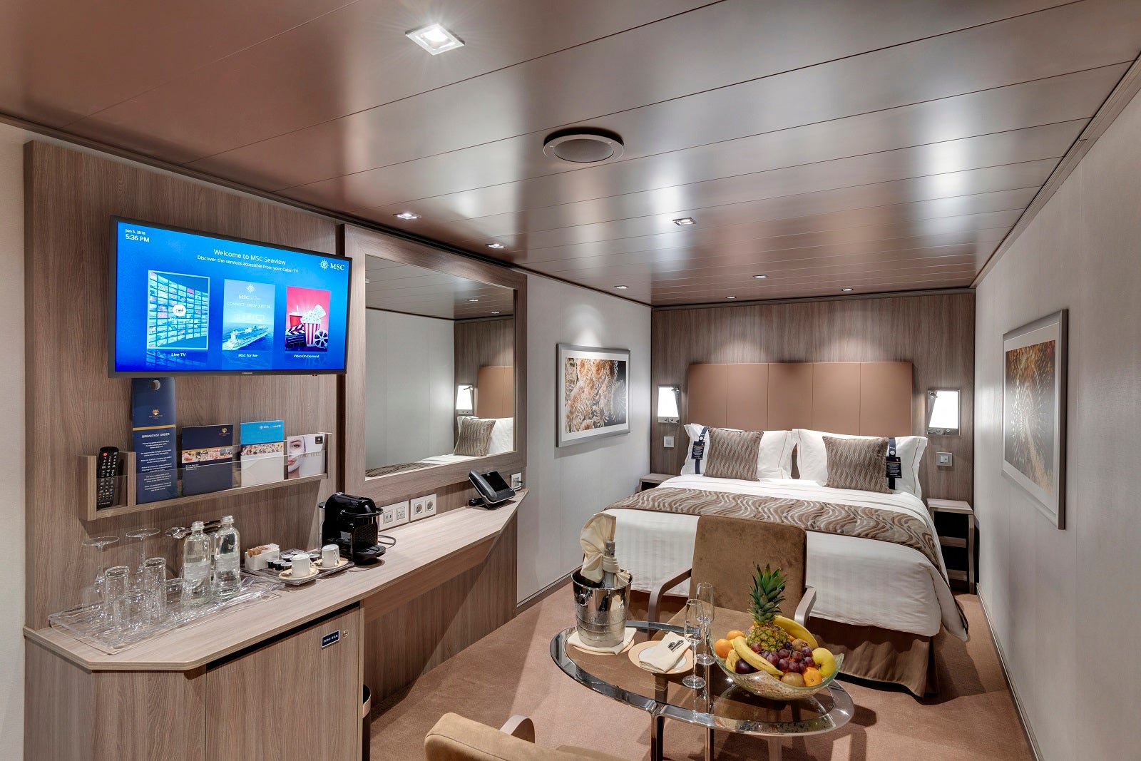 An interior cruise ship cabin decorated in neutral colors with a bed, desk, TV and mirror